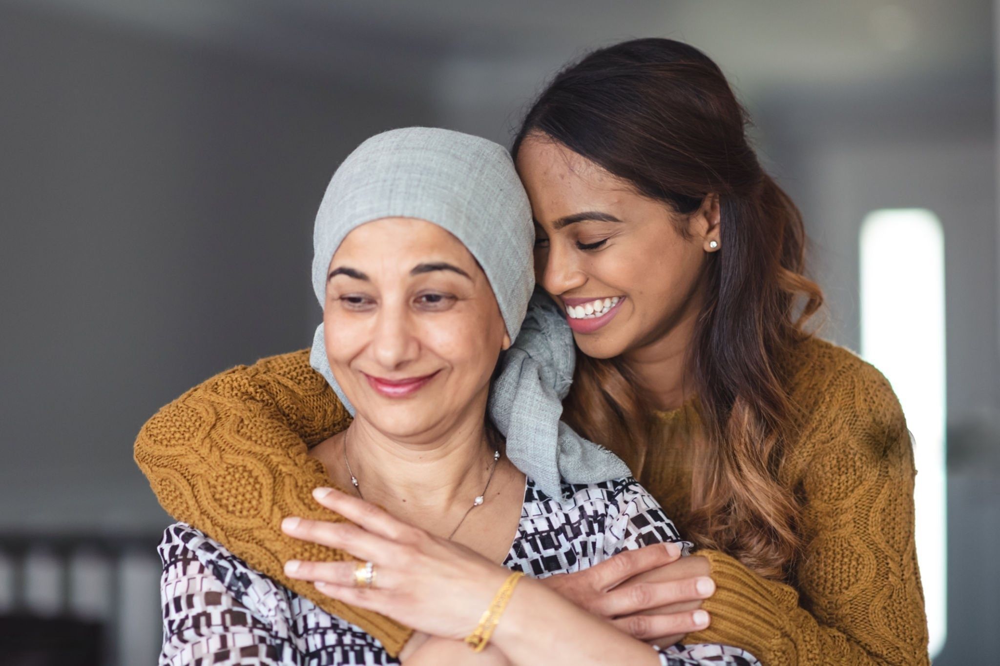 A smiling young woman hugging her mother, who wears a headscarf during cancer treatment